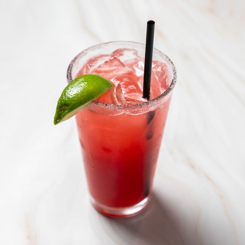 A vibrant red drink with ice, a black straw, and a lime wedge on a white marble surface.